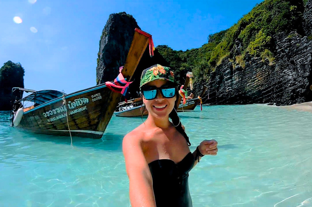 A woman taking a selfie in shallow turquoise water with tropical cliffs and longtail boats in the background in Thailand