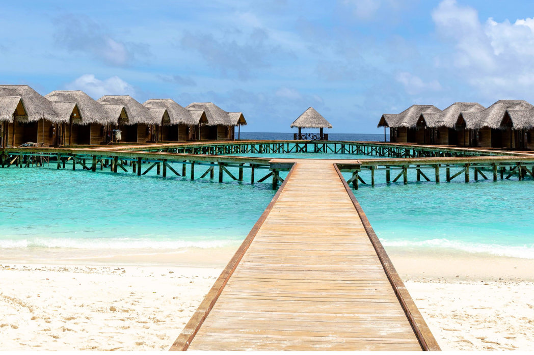 A wooden boardwalk pier extends over bright turquoise water toward overwater bungalows with thatched roofs on a white sandy beach under a blue sky.